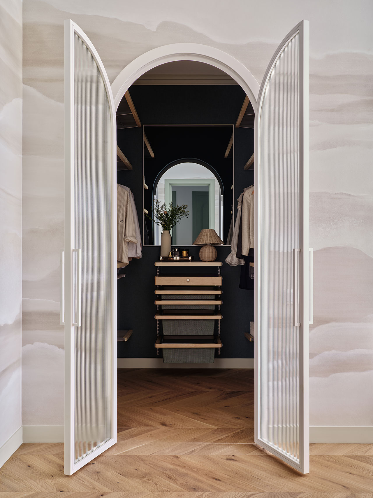 Open double doors reveal a walk-in closet with hanging clothes, wooden shelves, a round mirror, a vase with flowers, and a lamp on a dark accent wall.