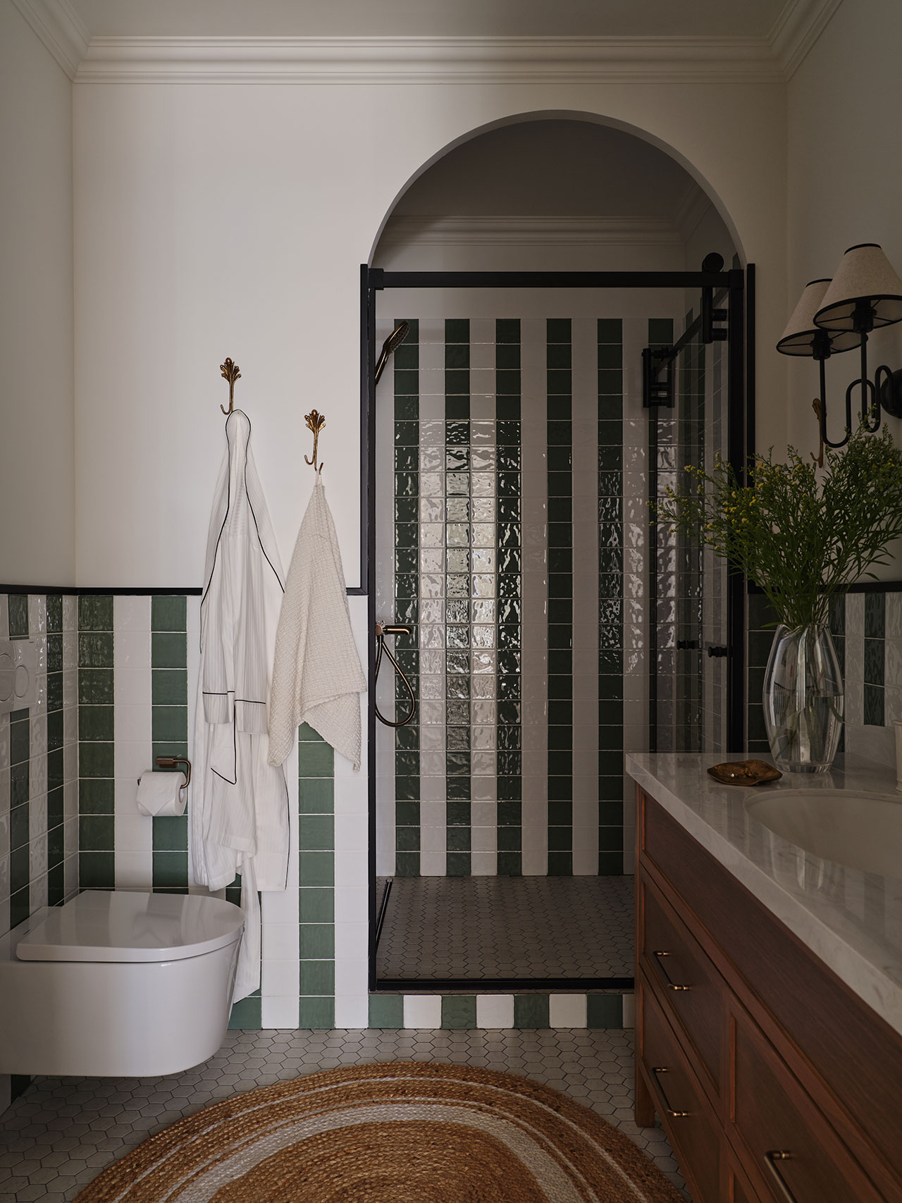 Bathroom with green and white vertical tile, glass shower enclosure, wall-mounted toilet, two robes on hooks, a vanity with drawers, and a vase with greenery.
