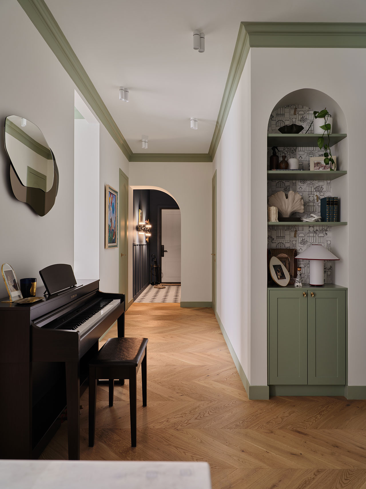 A hallway with light wood herringbone flooring, a green-trimmed arch, built-in shelves, and a black digital piano with a bench against the wall.