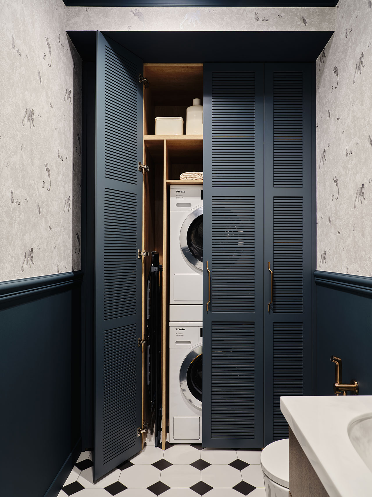 A stacked washer and dryer are enclosed in a navy blue louvered closet with shelves, located in a small room with patterned wallpaper and a black-and-white tile floor.