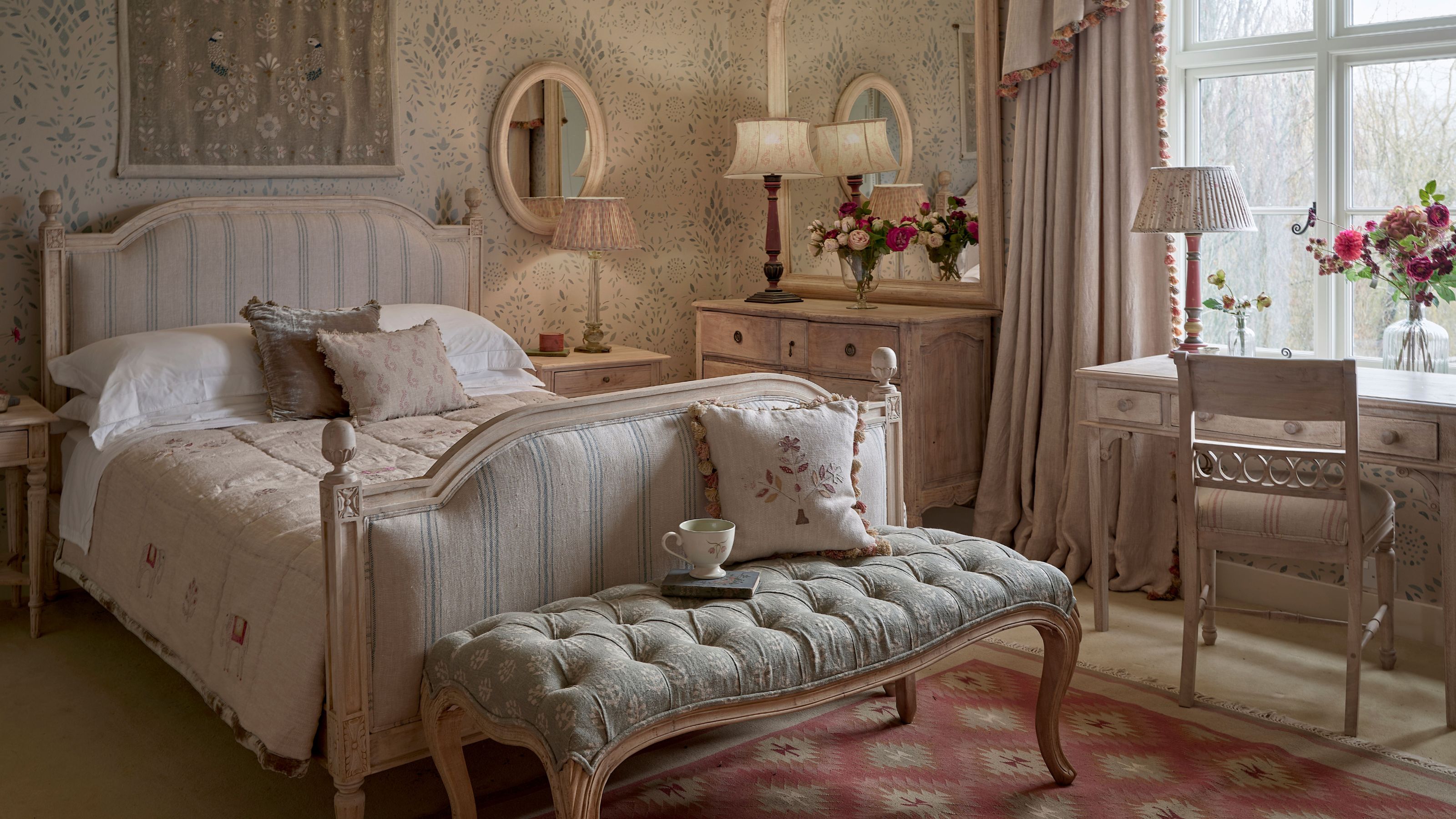 Vintage-style bedroom featuring a striped upholstered bed frame, a tufted bench on a pink rug, and a wooden writing desk by a window.