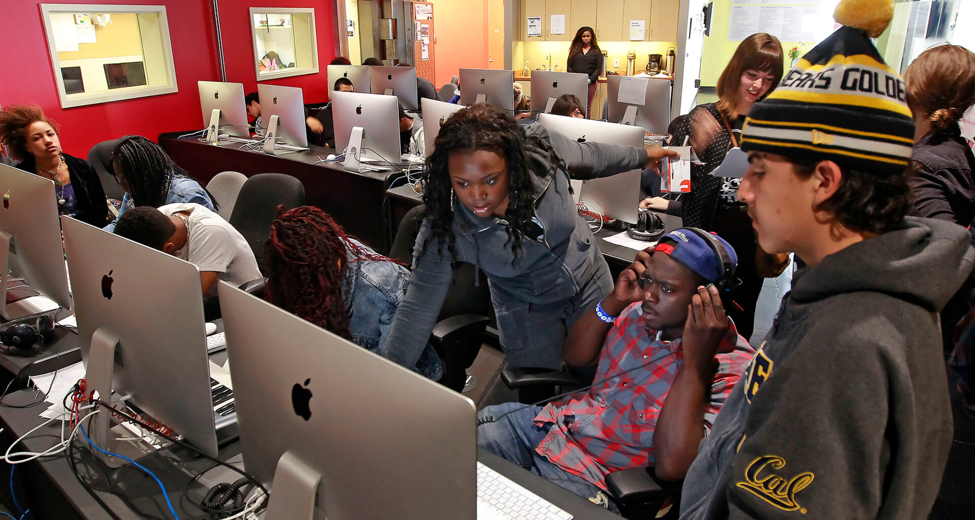 A young Black woman in blue denim helps a multiethnic group of students in front of a room of desktop computers.