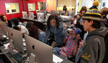 A young Black woman in blue denim helps a multiethnic group of students in front of a room of desktop computers.