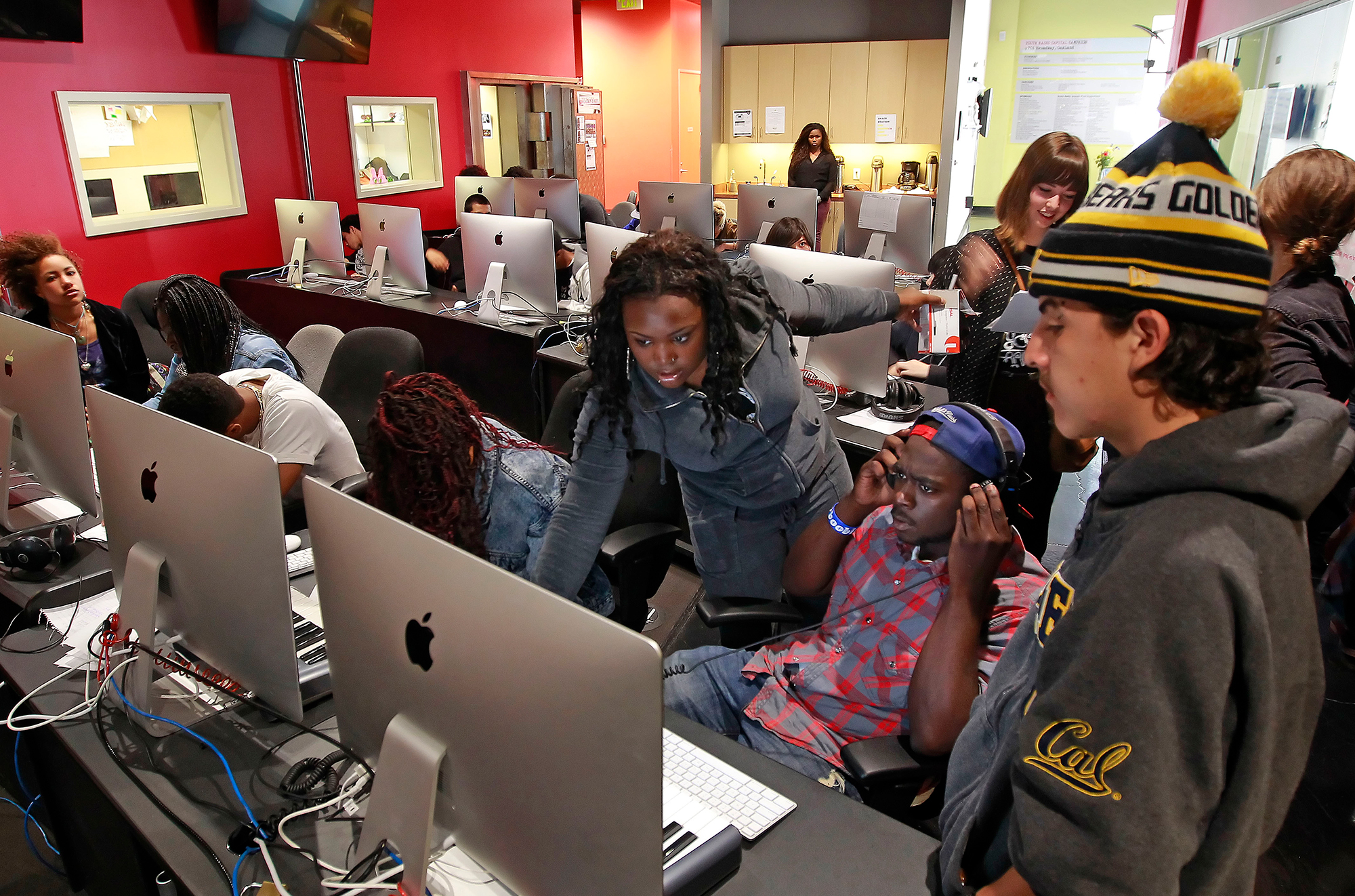 A young Black woman in blue denim helps a multiethnic group of students in front of a room of desktop computers.