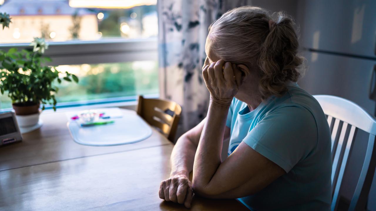 Older woman in blue shirt sitting at kitchen table, gazing pensively out window