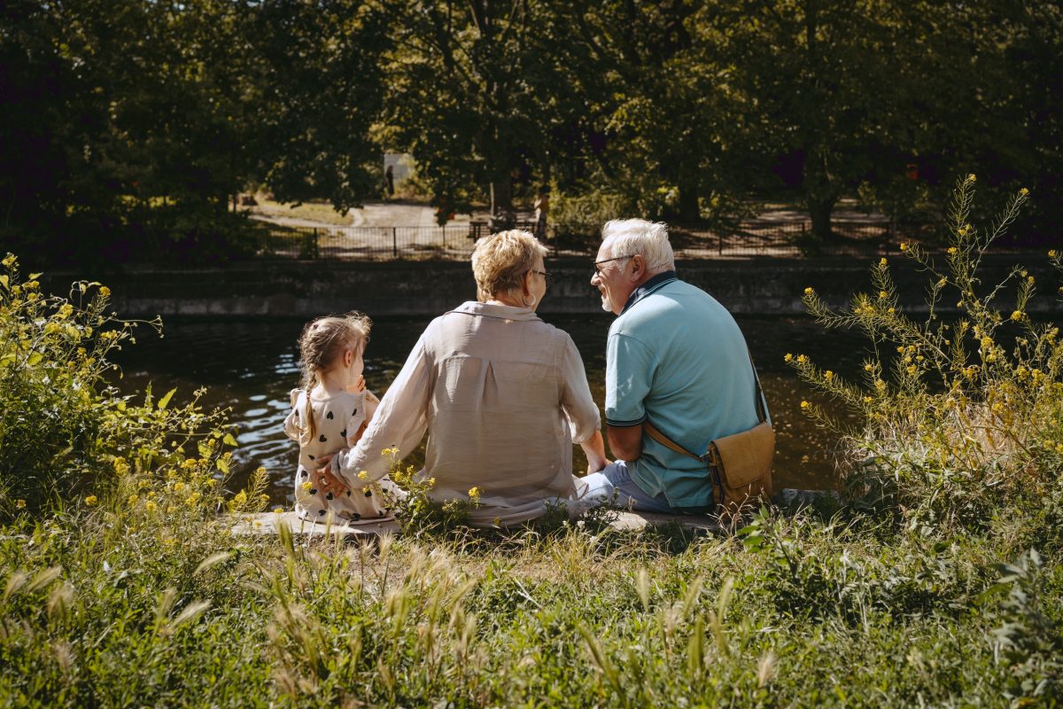 Rear view of grandparents sitting with granddaughter near pond at park - stock photo