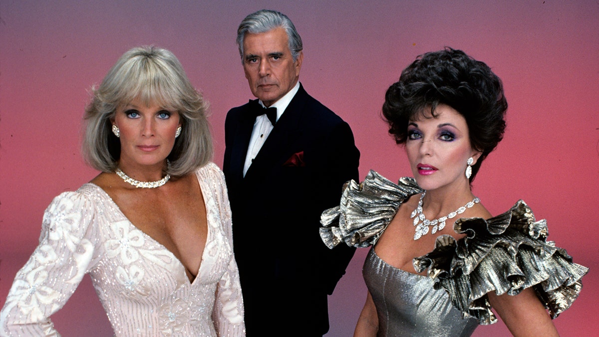 Linda Evans, John Forsythe, and Joan Collins standing together in 1980s fashion on Dynasty set