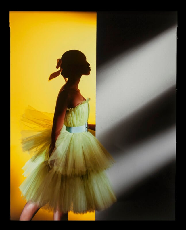 A person in a light green tulle dress and hair ribbon stands profile to a wall, illuminated by yellow light on one side and dramatic shadows on the other.
