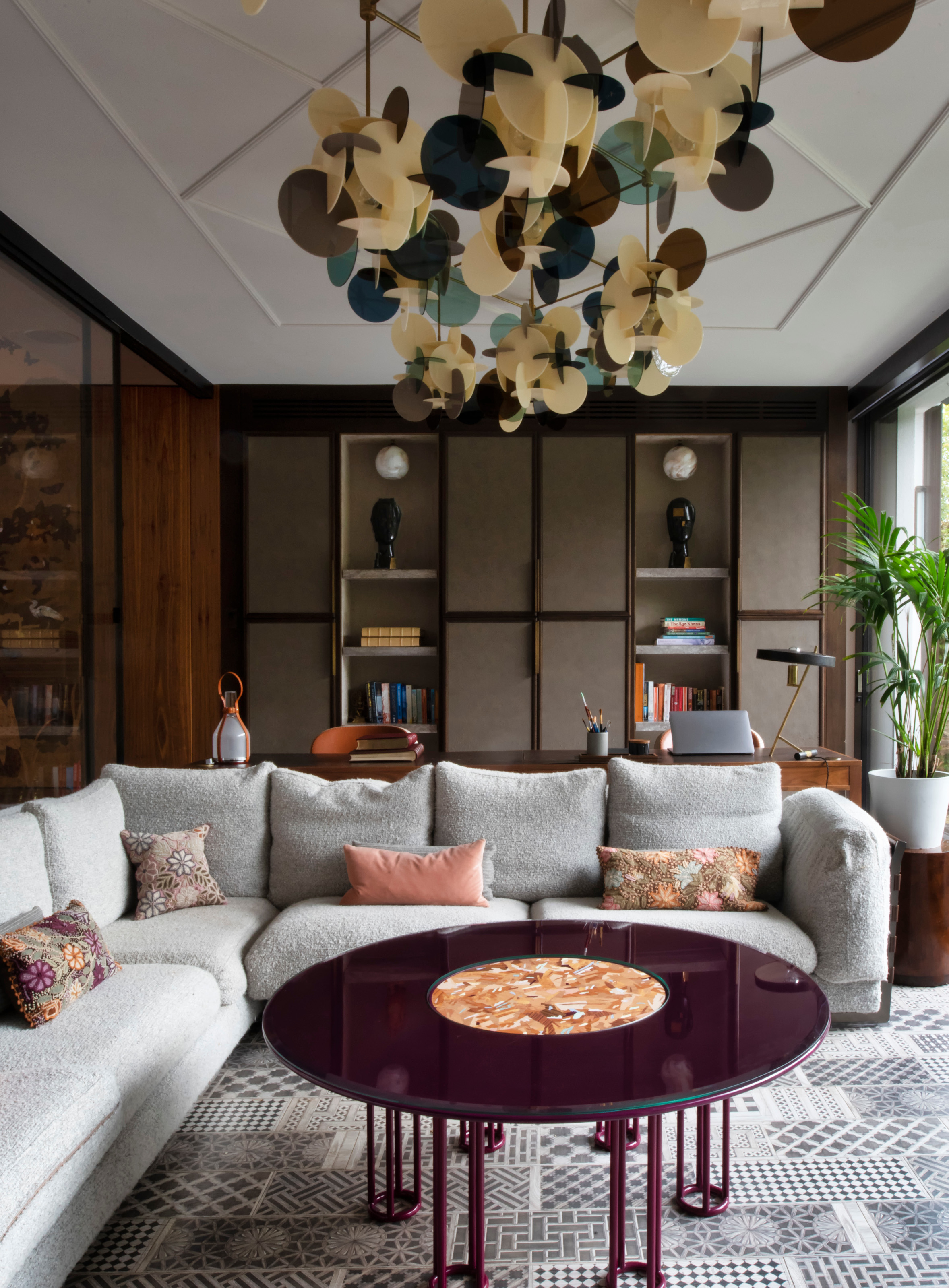 Living room with white panelled ceiling, grey corner sofa, burgundy gloss coffee table, geometric grey rug, and wooden desk in background against green fabric panelled joinery