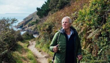 A senior man using hiking poles, on a hike on a coastal path along the coastline of Cornwall. He is looking away from the camera at the view.