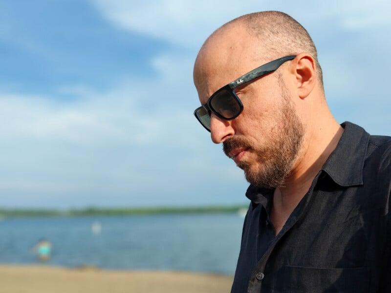 A man with a shaved head, beard, and sunglasses stands on a beach wearing a dark shirt. The background shows water, sky, and a distant person near the shoreline. The lighting is bright and natural.