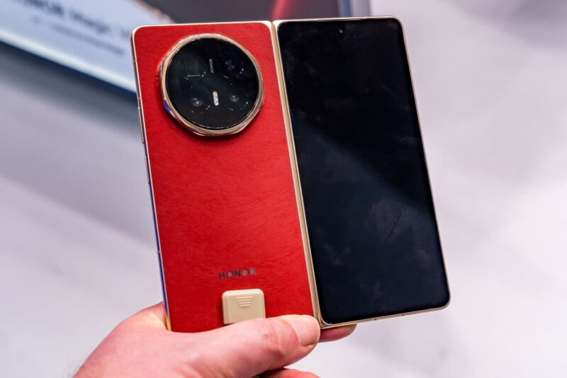 A hand holds an HONOR foldable smartphone with a red textured back, a large circular camera module, and a gold frame. The phone is partially unfolded, revealing its black screen.