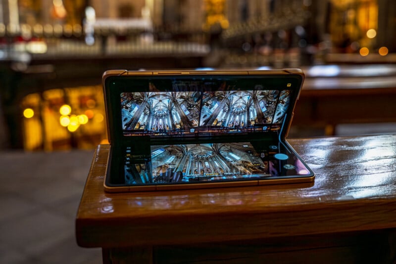 A foldable smartphone sits open on a wooden surface, displaying a detailed photo of an ornate, vaulted church ceiling. The background features a softly blurred interior of the same church with warm lighting.