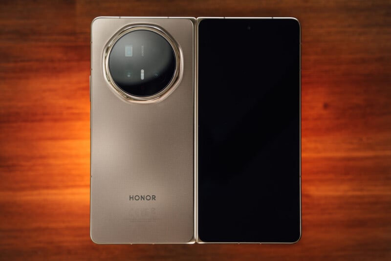 A foldable HONOR smartphone is shown open on a wooden surface, displaying its large black screen on the right and the textured back with a circular camera module on the left.