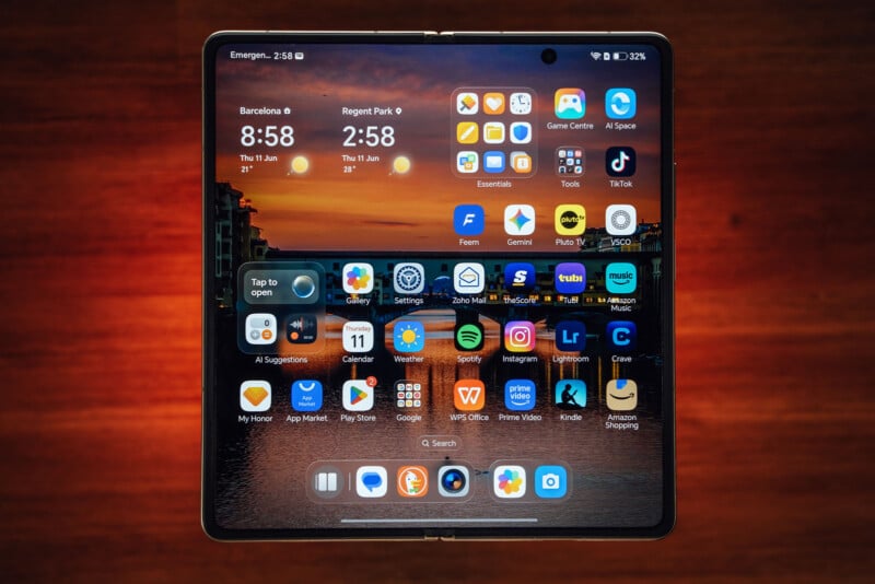 A foldable smartphone with its screen open, displaying a sunset wallpaper and a grid of various app icons, sits on a wooden surface. The device shows clocks for Barcelona and Regent Park at the top.