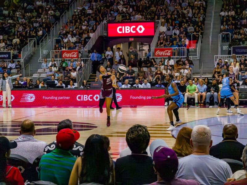 A basketball player in a maroon jersey runs and catches a ball midair as a defender in a blue jersey approaches, with a packed crowd watching in an indoor arena. Bright sponsor signs are visible in the background.