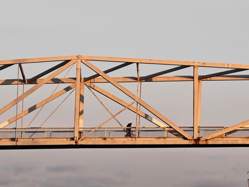 A person rides a bicycle across a metal bridge with geometric beams at sunset, silhouetted against a pale sky.