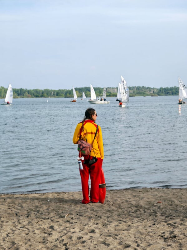 A lifeguard in a yellow shirt and red pants stands on a sandy beach, facing a lake with several small sailboats in the water. The weather is clear, and trees line the far shore.