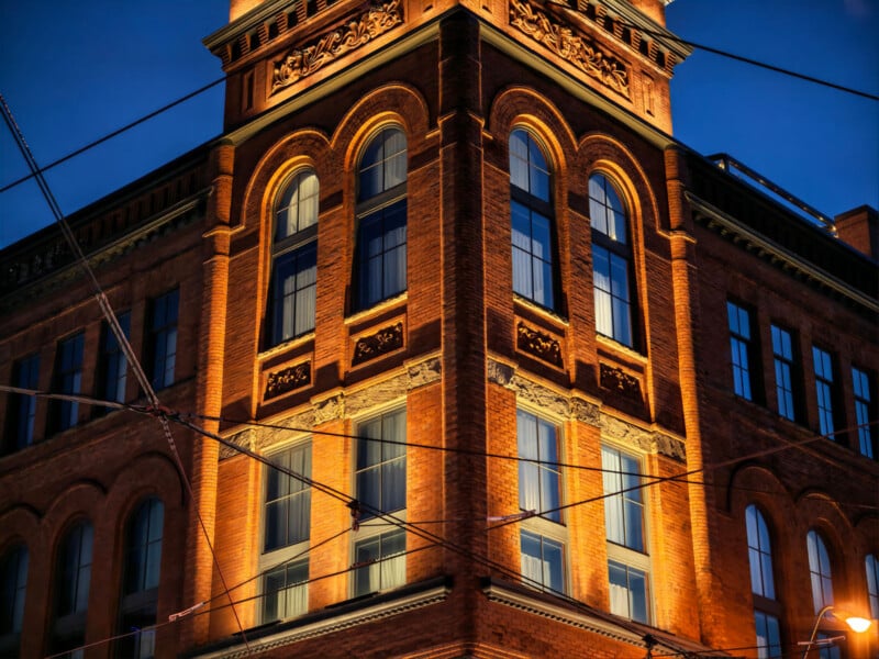 A red brick building with large arched windows is illuminated at night against a deep blue sky, with power lines crossing in front of the structure.