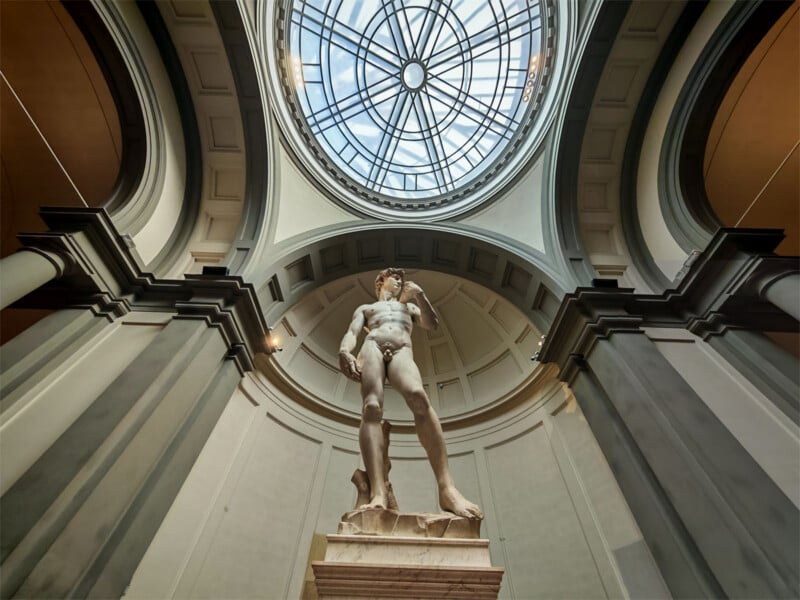 Low-angle view of Michelangelo’s statue of David beneath a large domed glass skylight in a grand, arched hall, emphasizing the sculpture’s height and architectural details.