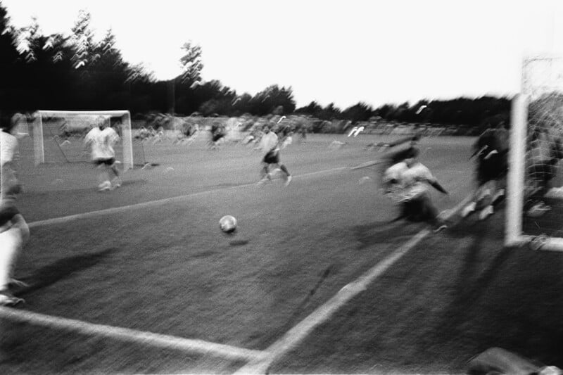 A black-and-white, motion-blurred photo of a soccer game shows players near the goal as a ball moves toward the net, with spectators and another goal visible in the background.