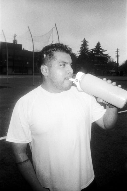 A man in a white shirt drinks from a large water bottle on an outdoor sports field, with netting and trees visible in the background. The image is in black and white.