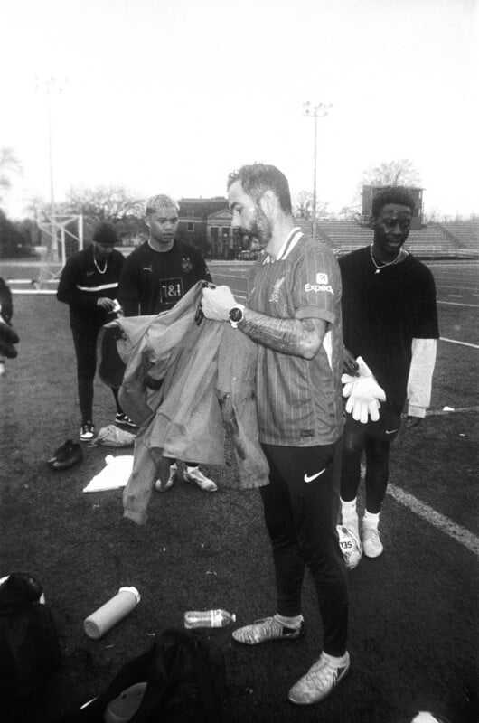 A group of young men stand on a sports field. One man in front holds a jacket and a pair of goalkeeper gloves, while others stand nearby with sports gear scattered on the ground. The setting appears casual and relaxed.