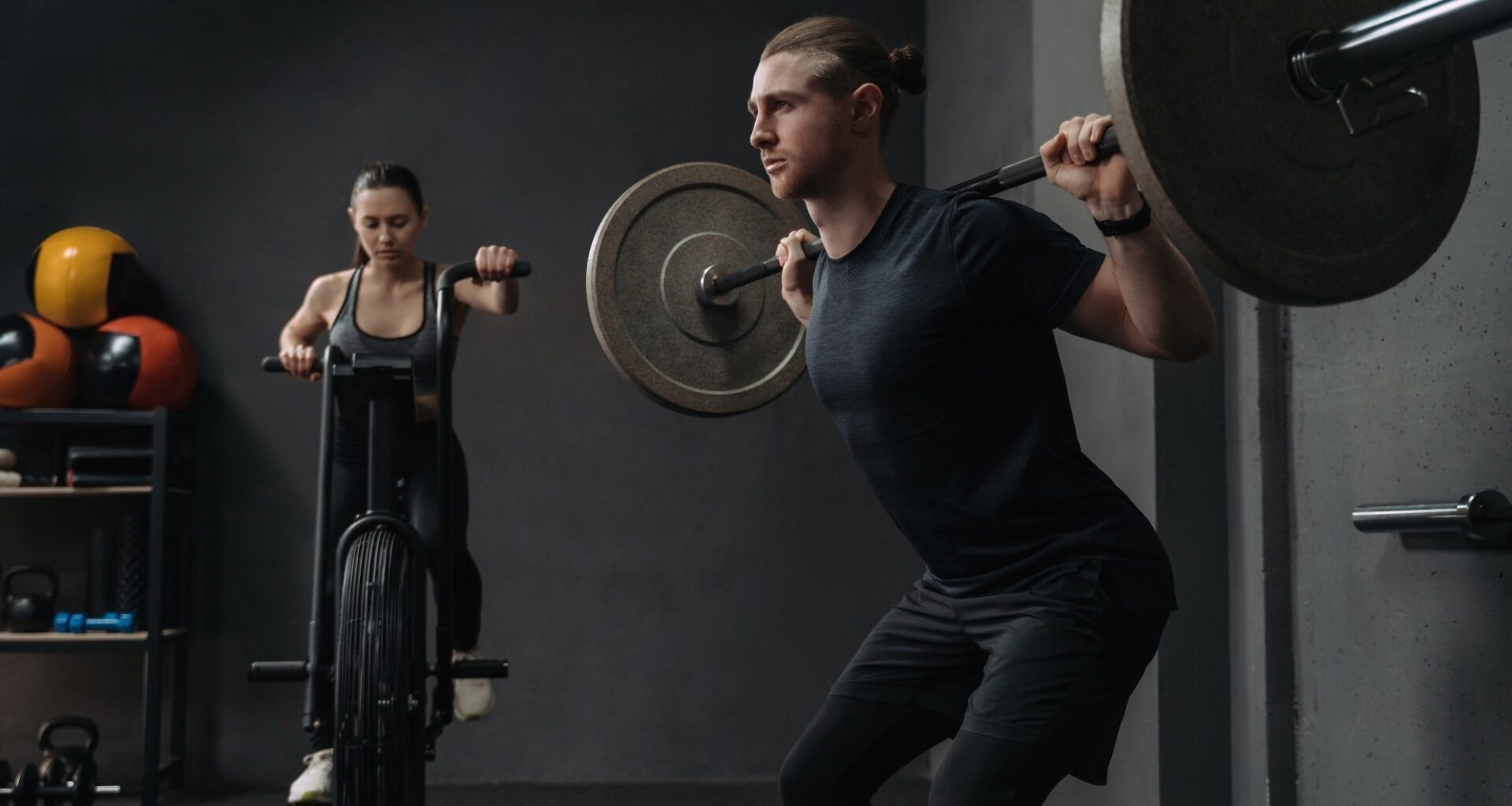 Fit woman is working out on exercise bike, while muscular man training with barbell.