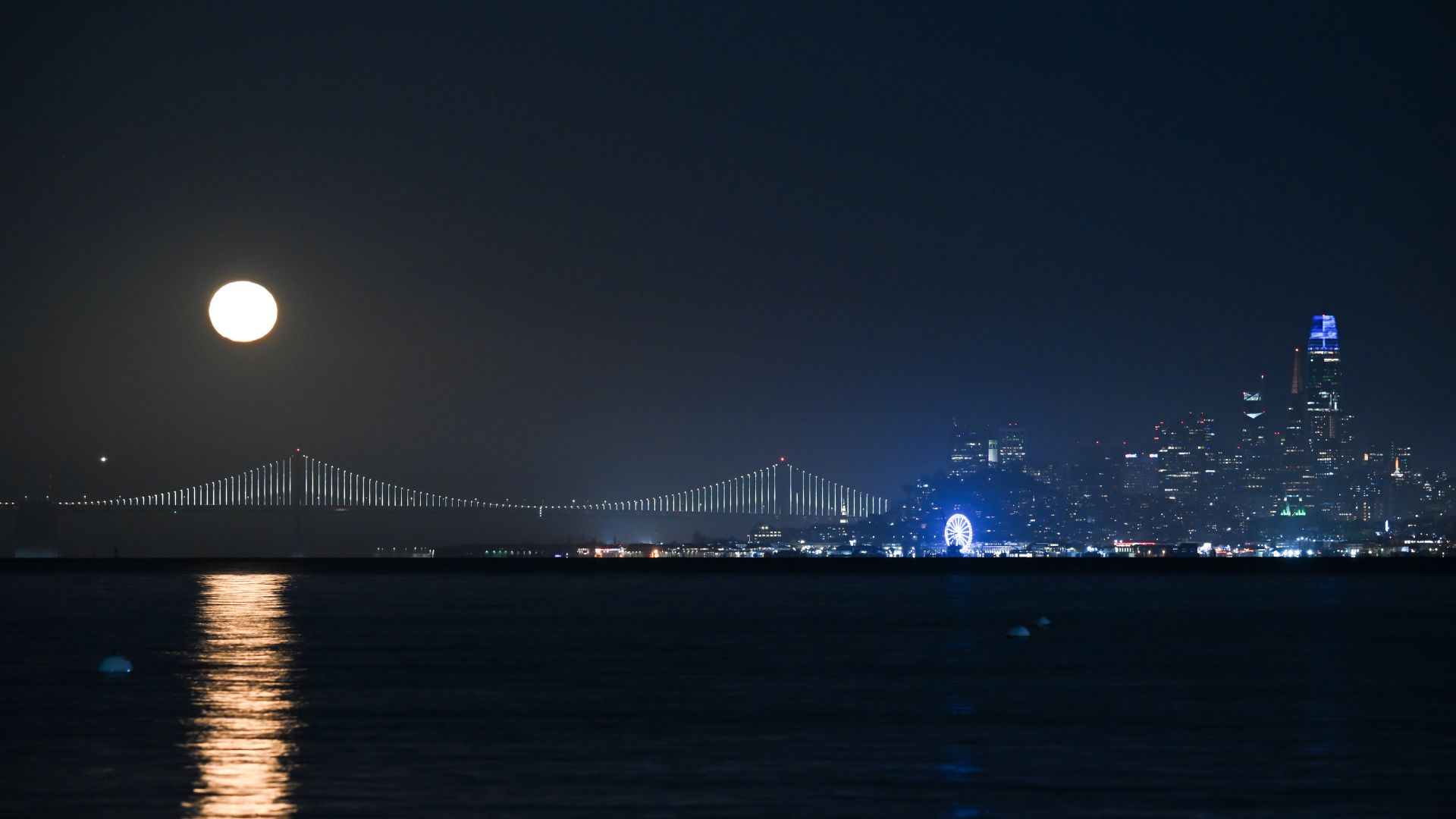 A full moon rises in a night sky above an illuminated suspension bridge, as a city skyline towers to the right. A body of water reflects the moonlight in the foreground.