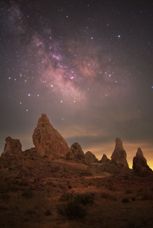 Tall rocky spires rise from a desert landscape under a night sky filled with stars and the glowing colors of the Milky Way, with soft clouds near the horizon.