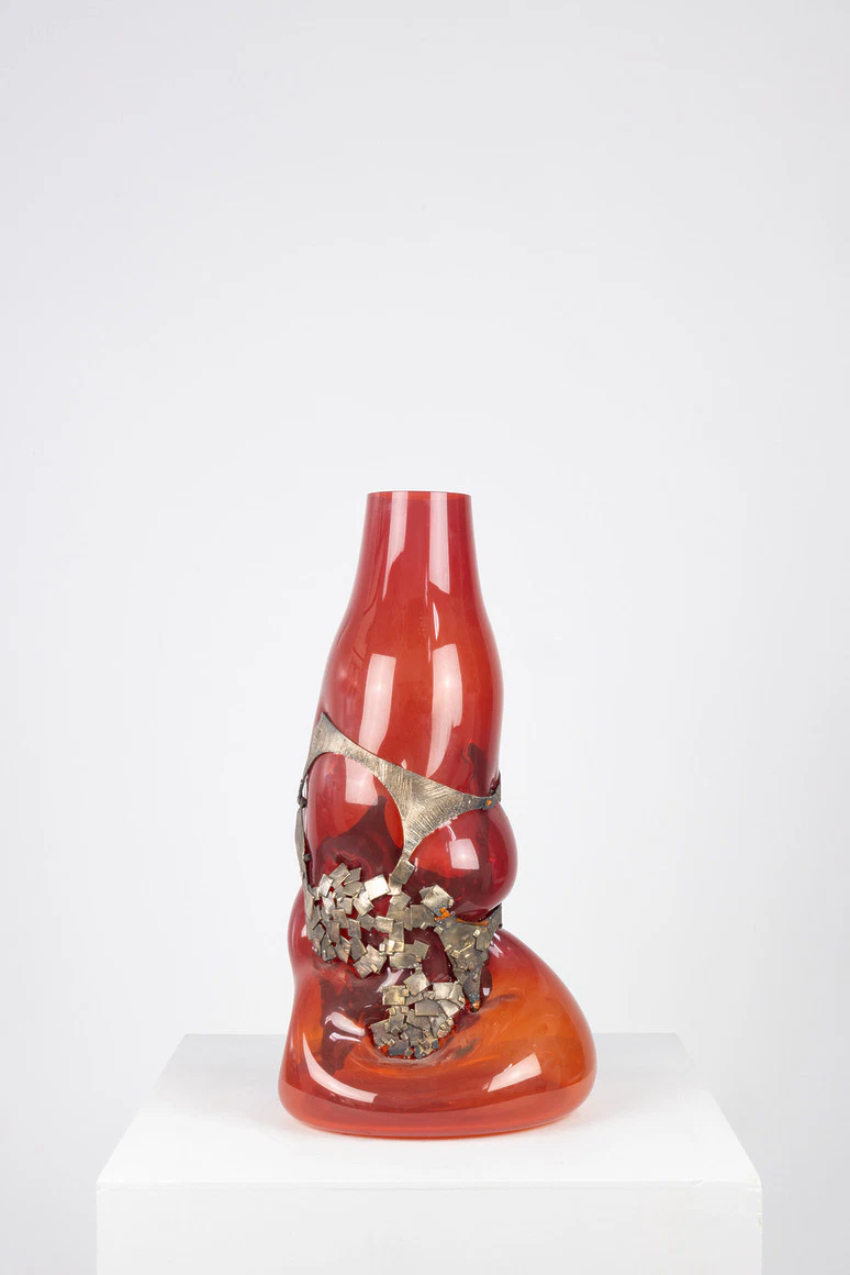 Abstract red glass sculpture with metallic details displayed on a white pedestal against a plain background.