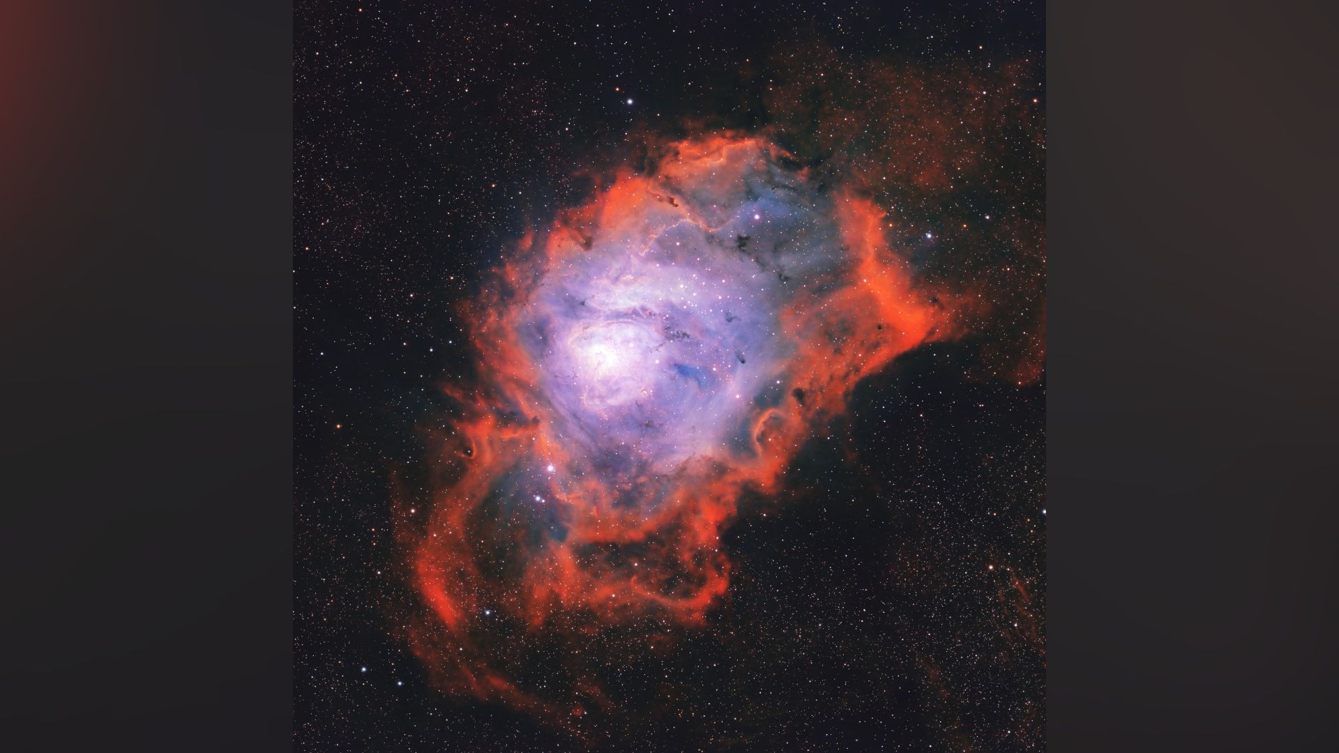 A nebula is photographed glowing in deep space among a sea of stars. It features a blue glowing core reminiscent of a lagoon, surrounded by red, flame-like clouds.