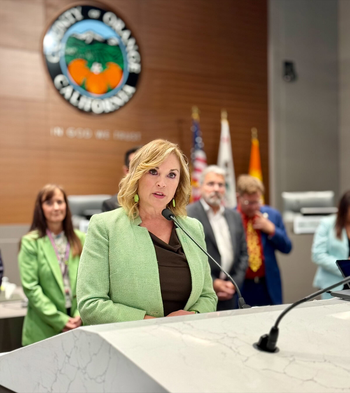 A woman speaks in a courtroom. She has blond hair and wears a green blazer.