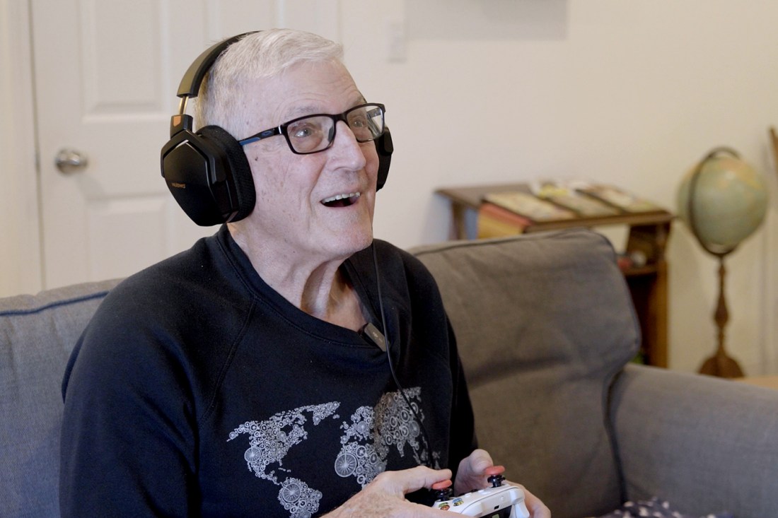 An older man playing a video game while smiling, wearing glasses and over the ear headphones on and holding a controller in hand. 