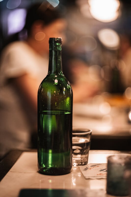 A green glass bottle filled with water stands on a table next to a clear drinking glass, with a blurred background of people and warm lighting.
