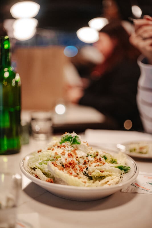 A bowl of Caesar salad topped with croutons and cheese sits on a table in a restaurant, with a green bottle and blurred people in the background.