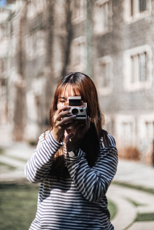 A person with long hair wearing a striped shirt is standing outdoors and holding a camera up to their face, taking a photo. The background shows a building in soft focus.