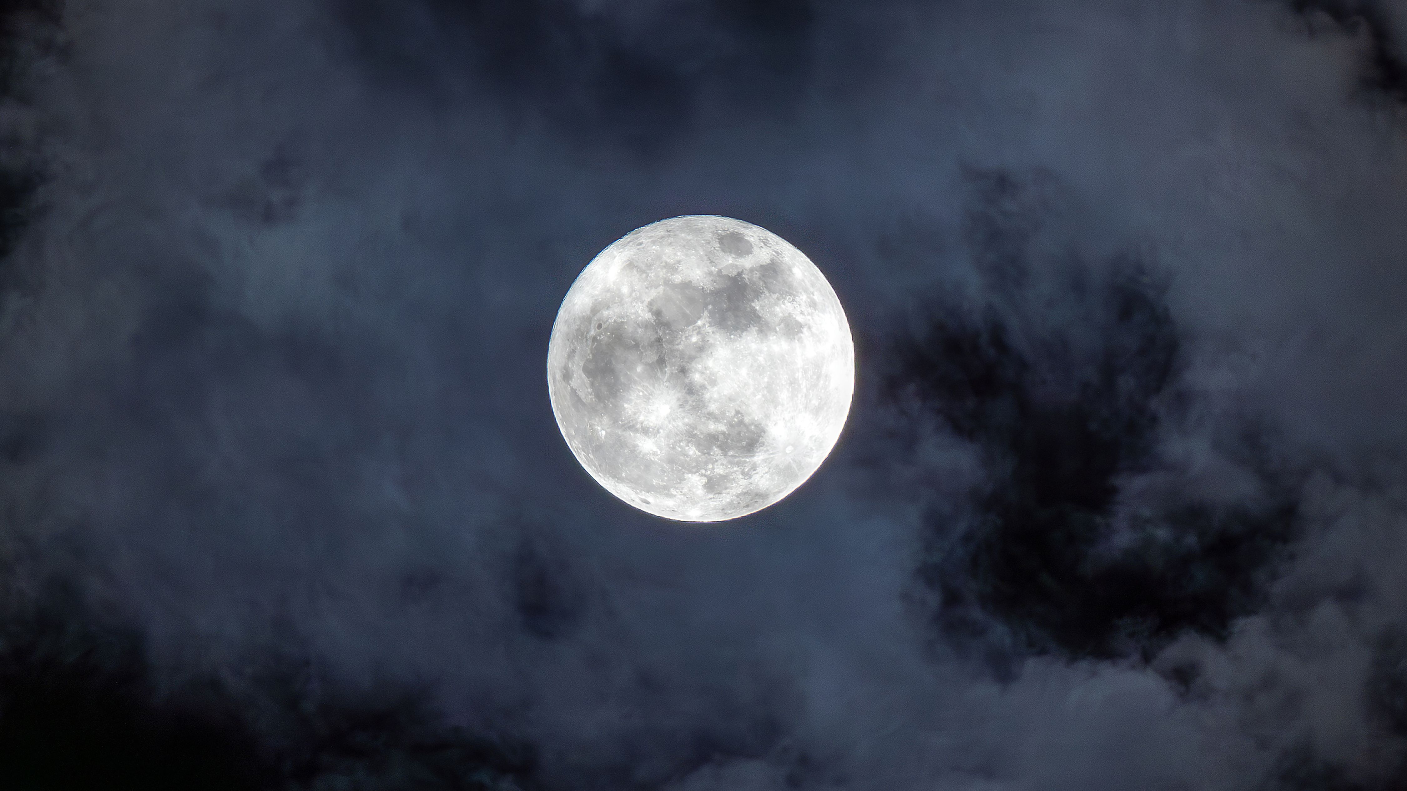 A silver full moon shines in a cloudy sky, as lunar seas darken its surface.