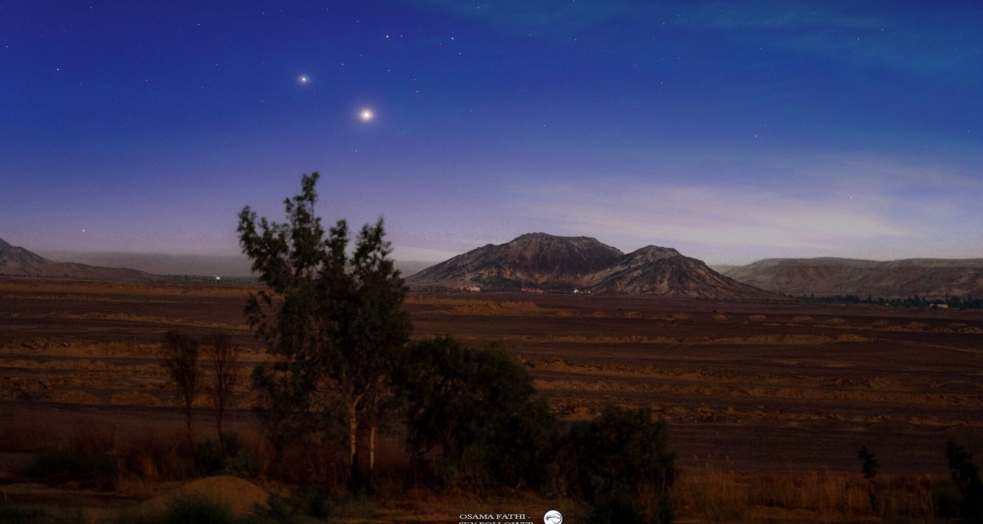 Two bright star-like planets are photographed shining side by side above mountains in a tranquil plain.