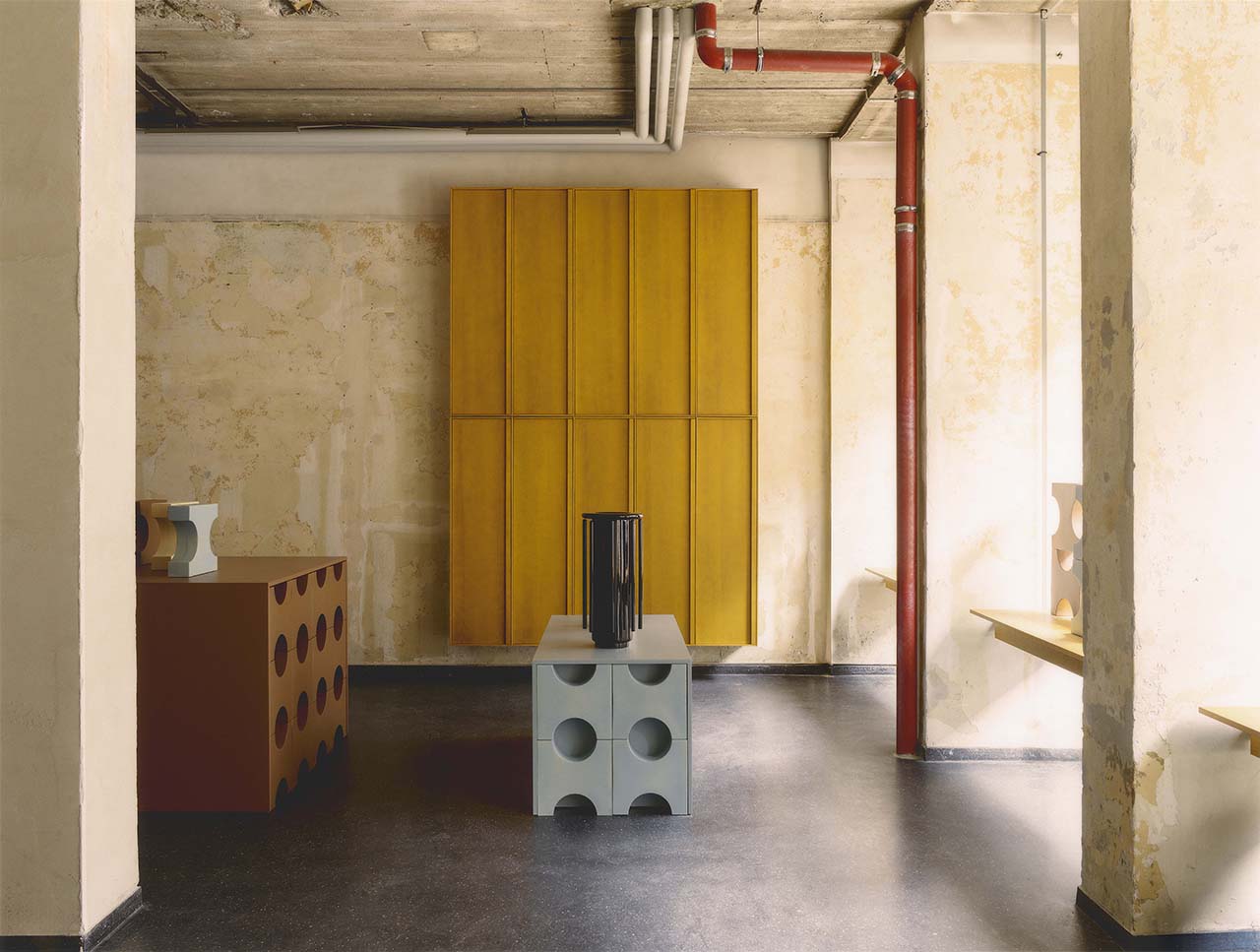 Minimalist interior with exposed concrete ceiling and pipes, mustard yellow wall panels, and geometric furniture featuring cube shapes with circular cutouts.