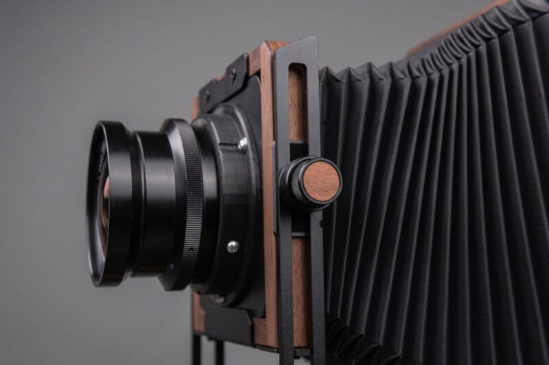 A close-up view of a large format camera, highlighting the black accordion-style bellows, metal lens, and wooden accents against a neutral background.
