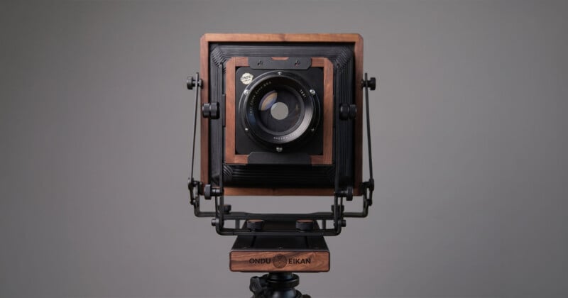 A large format wooden field camera with a bellows, metal supports, and a lens mounted at the center, set against a plain gray background. The camera is labeled "ONDU EIKAN.