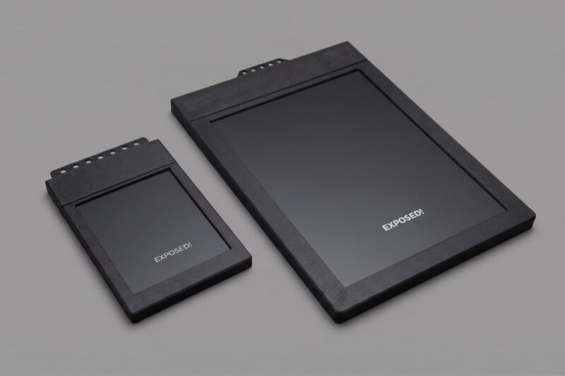 Two rectangular black film holders, one smaller and one larger, are laying on a gray surface. Both have the word "EXPOSED!" written in white on their fronts.