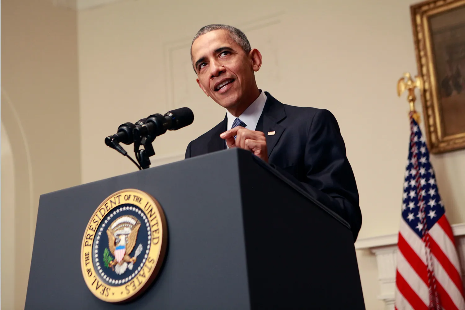 A low-angle, medium shot of President Obama in a dark suit, white shirt, and blue tie speaking at a dark grey podium. The podium features the circular Seal of the President of the United States on the front. He is gesturing with his left hand while looking slightly to the right of the camera. To his right, an American flag on a flagpole is visible in the background against a light-colored wall.