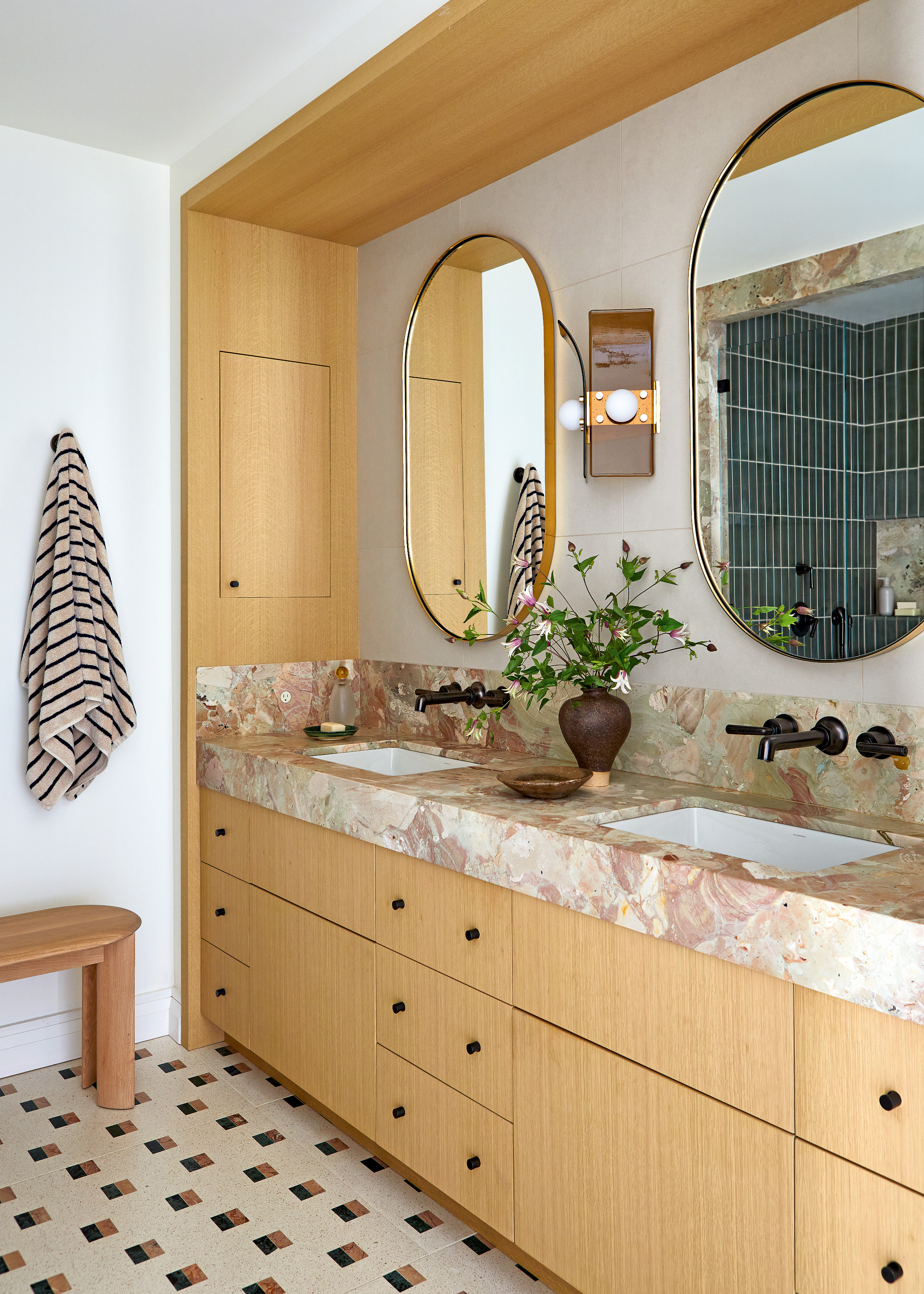 Bathroom with wood cabinetry, pink marble countertop, black hardware and oval mirrors