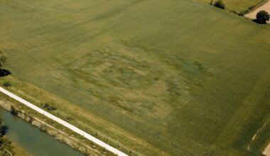 Aerial view of a large green field with a faint, lighter rectangular patch of different shades in the grass, possibly forming a subtle pattern or design. A road and a canal run along the edge of the field.