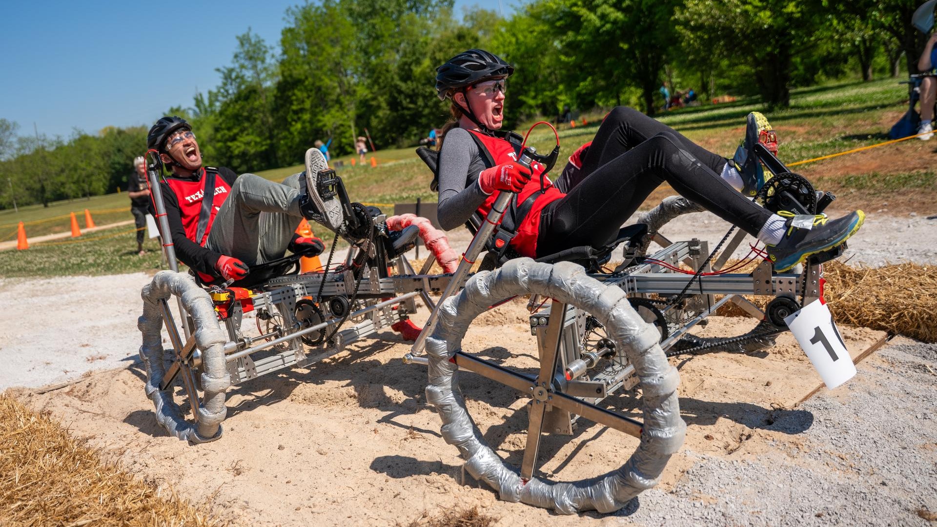 two people in bike helmets scream while pedaling a tandem-seat, four-wheeled vehicle on a dirt path