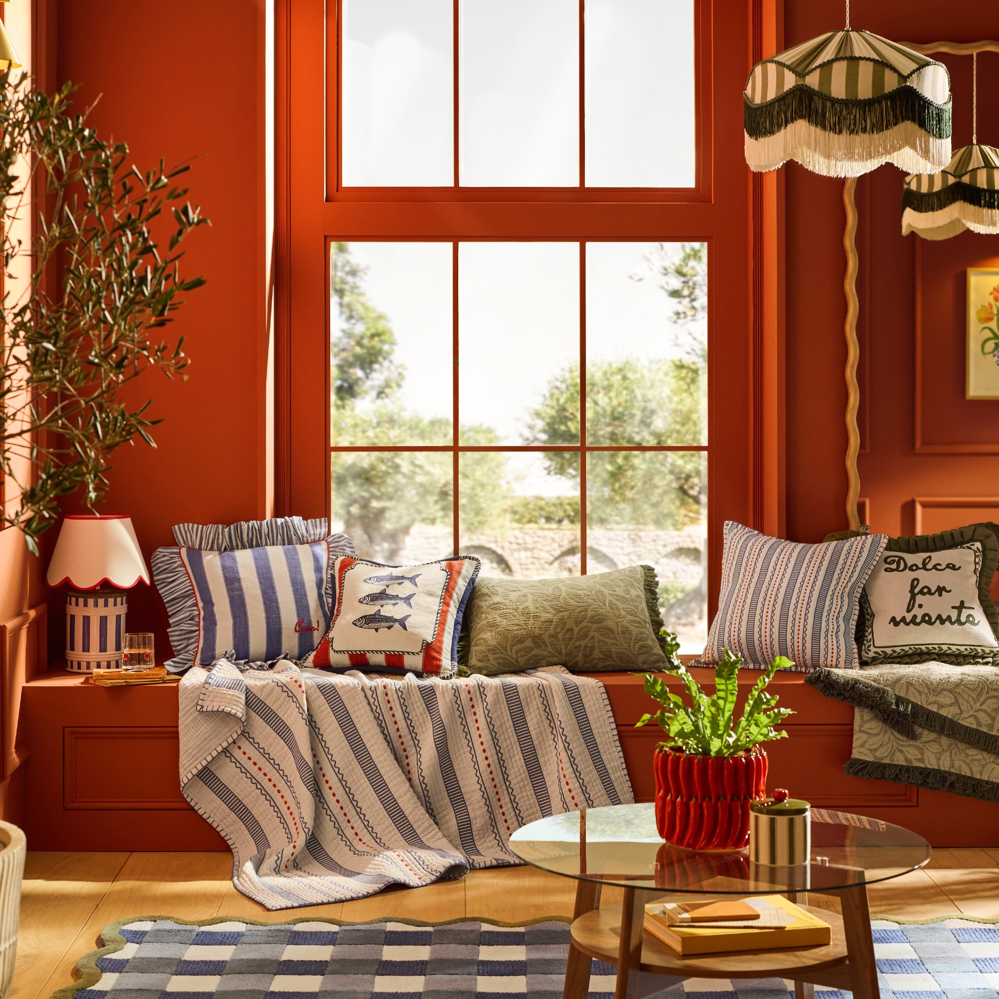 A red-painted living room with a window seat decorated with pieces from Lia Gold's collection for Next including striped cushions and a matching throw and striped, fringed pendants