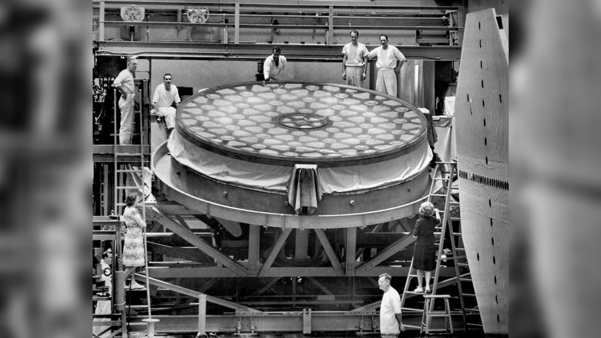 A black and white photo of a giant flat mirror and people in white outfits standing around it.
