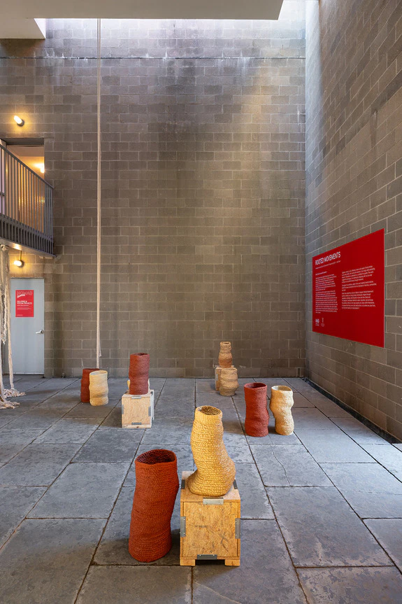 Art installation featuring woven basket sculptures arranged on wooden blocks in an industrial gallery space with informational signs.